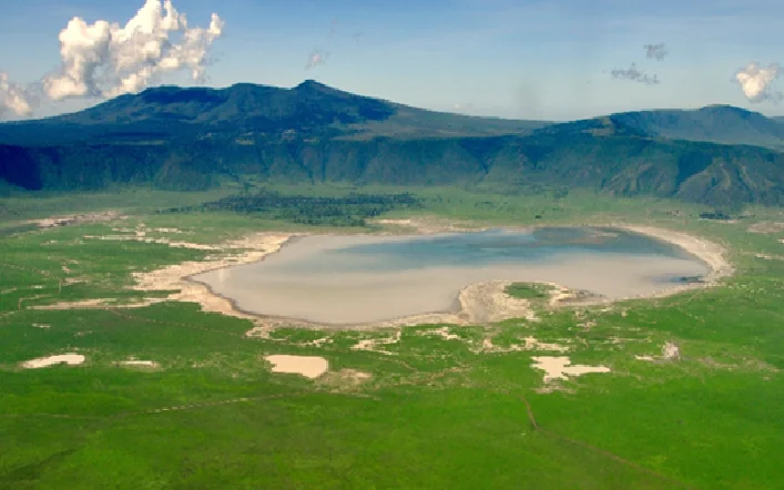 Aerial view of a pale blue crater lake surrounded by green plains and distant mountains on a sunny day, with a clear sky.