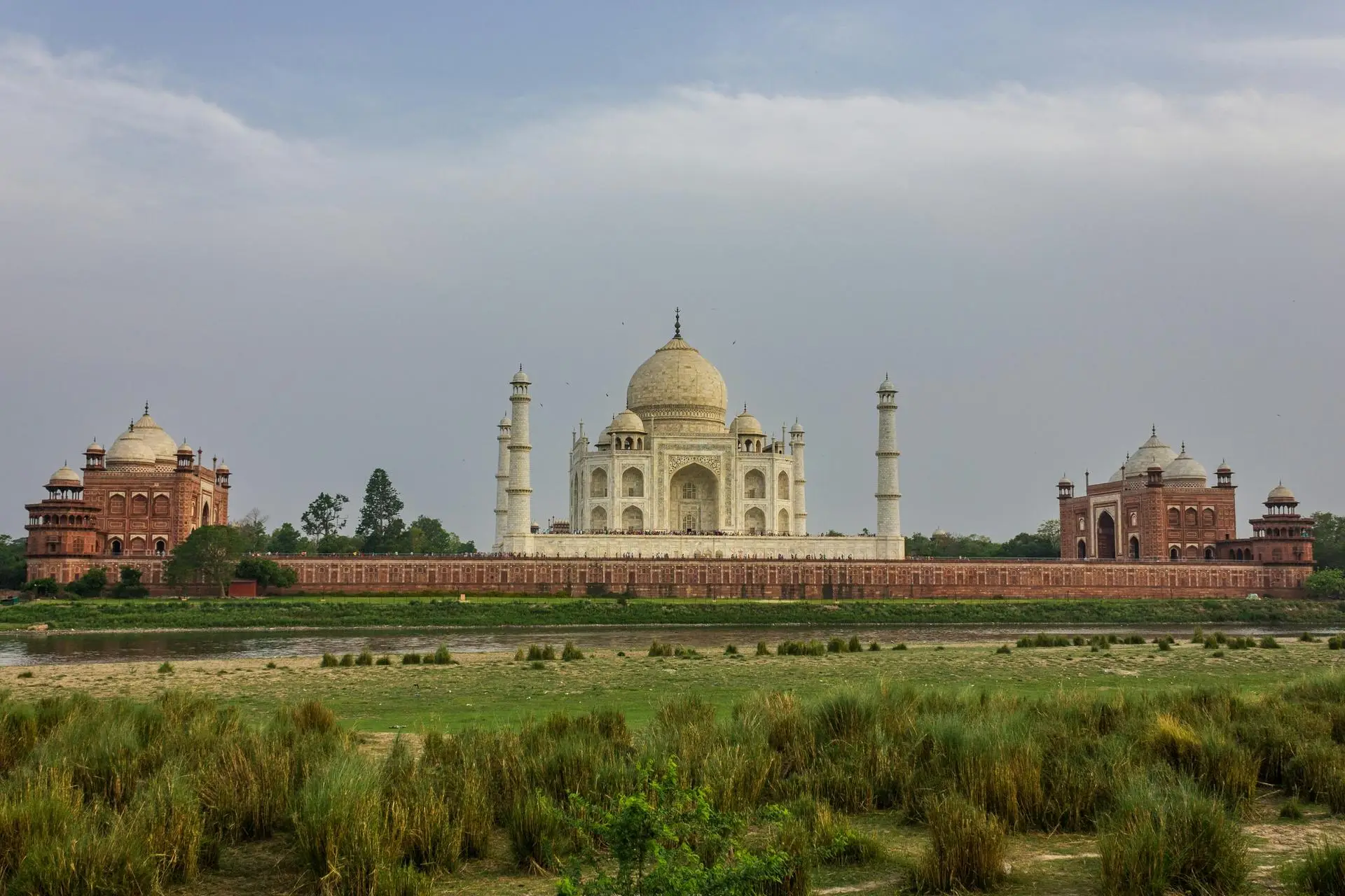 Taj Mahal centered in the image, white marble mausoleum with a large dome and four surrounding minarets, flanked by red sandstone buildings; river and greenery in foreground.