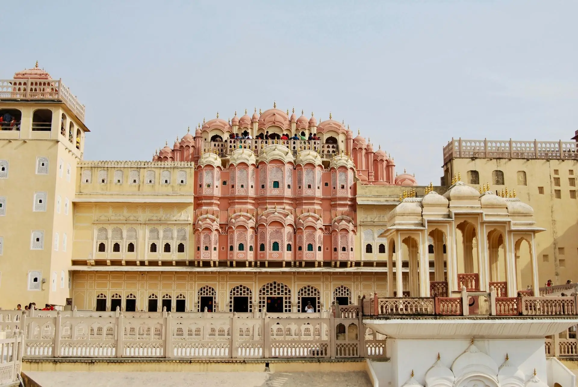 Front view of a large pink Indian palace with multiple domes, arches, and balconies along a cream-colored wall; visitors on the upper levels visible.